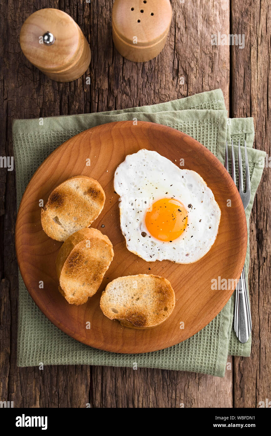 Una fresca uovo fritto sunny side up con baguette tostate fette sul lato servite sul piatto di legno, fotografato overhead (messa a fuoco selettiva) Foto Stock