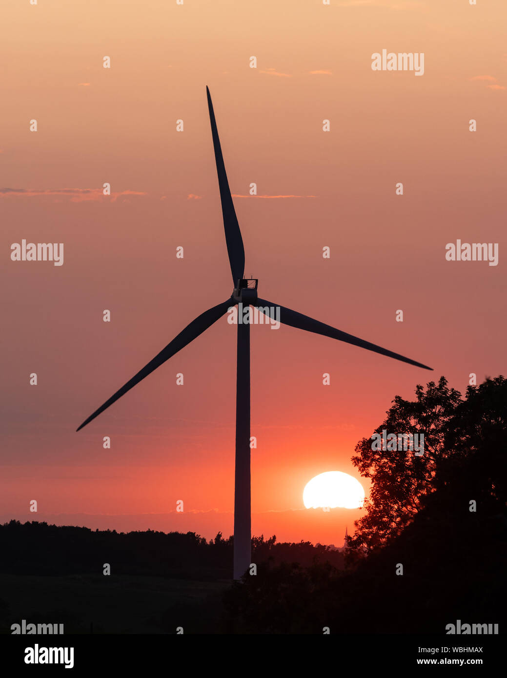 Crick, Northamptonshire, Regno Unito: Il sole, parzialmente oscurato dalle nuvole, tramonta in un cielo arancione dietro una turbina e un albero vento silhouette. Foto Stock