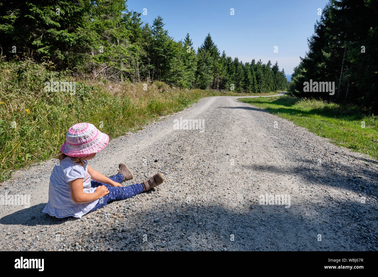 Vista posteriore di un 3-4 anno vecchia ragazza bambino in estate abbigliamento seduto su una strada di ghiaia in un bellissimo bosco su un tour escursionistico e non vuole camminare un Foto Stock