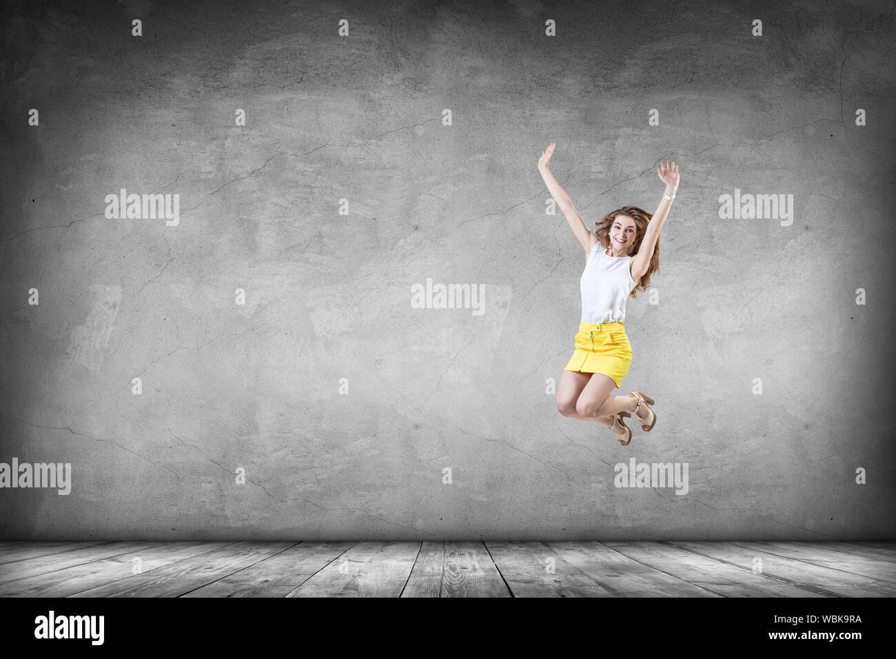 Felice giovane donna nel mantello giallo salta verso l'alto. Oltre il muro grigio Sfondo. Foto Stock