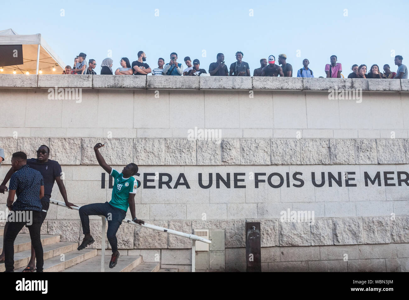 Lione, Francia - 14 luglio 2019: giovane nero French uomo seduto sulle scale prendendo un selfie con il suo smartphone di fronte ad una folla sulla riva del fiume del Foto Stock