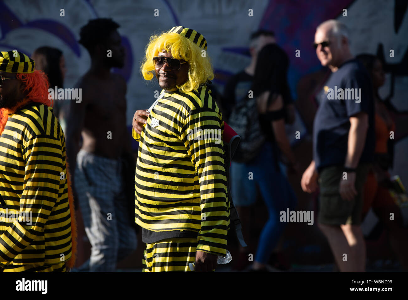 Un uomo al carnevale di Notting Hill che indossa un nero e giallo e del costume che indossa una parrucca di colore giallo mentre camminando attraverso una folla. Foto Stock