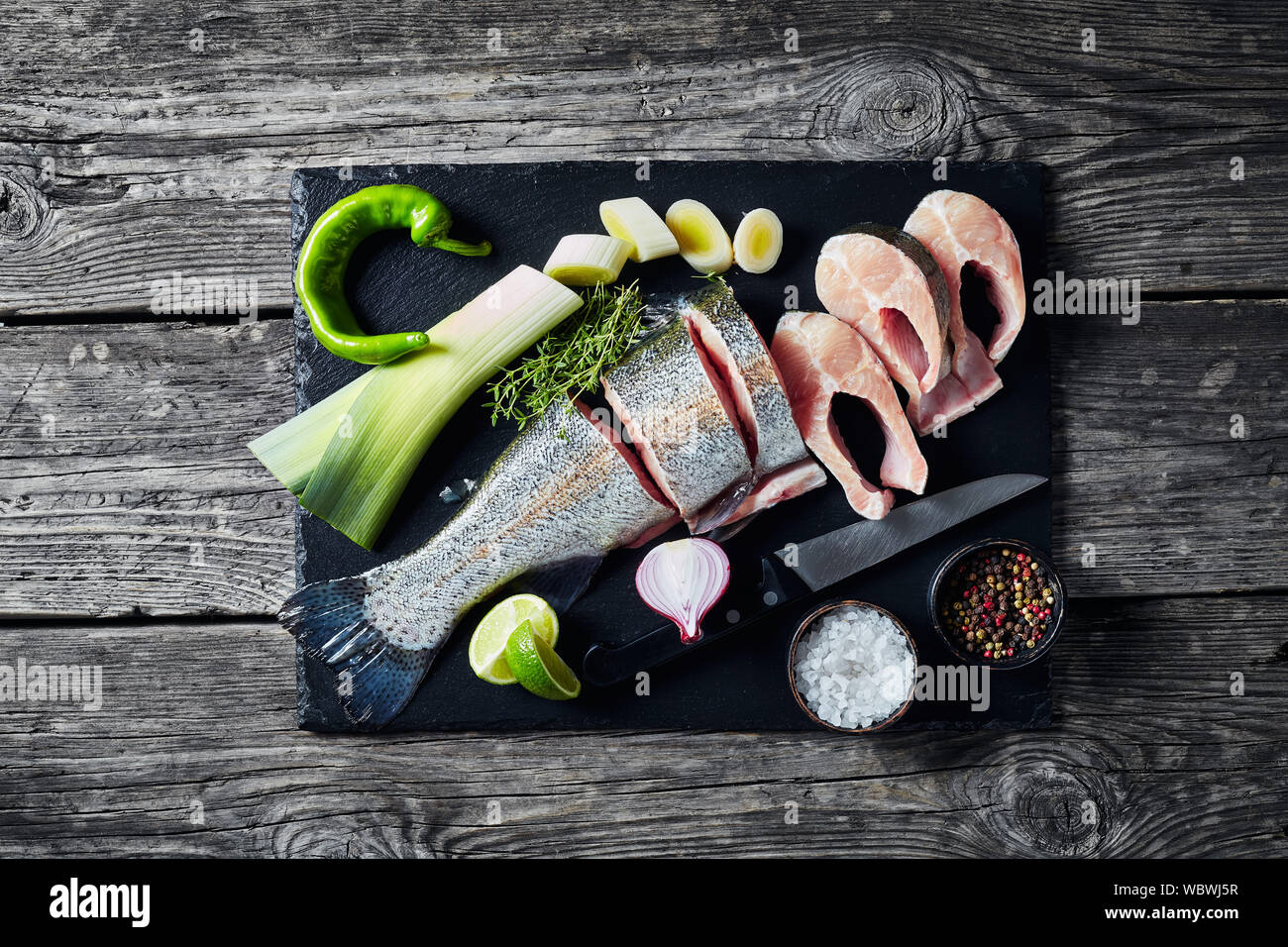 Fette di materie di trote fresche con spicchi di lime, porro, timo, sale e spezie su una pietra nera tagliere su una vecchia tavola in legno rustico, vista orizzontale f Foto Stock