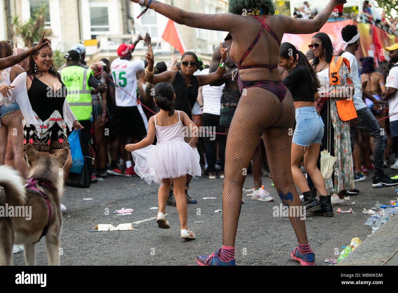 Una piccola ragazza in un abito bianco ballando con il carnevale di Notting Hill ballerini in mezzo alla strada. Foto Stock