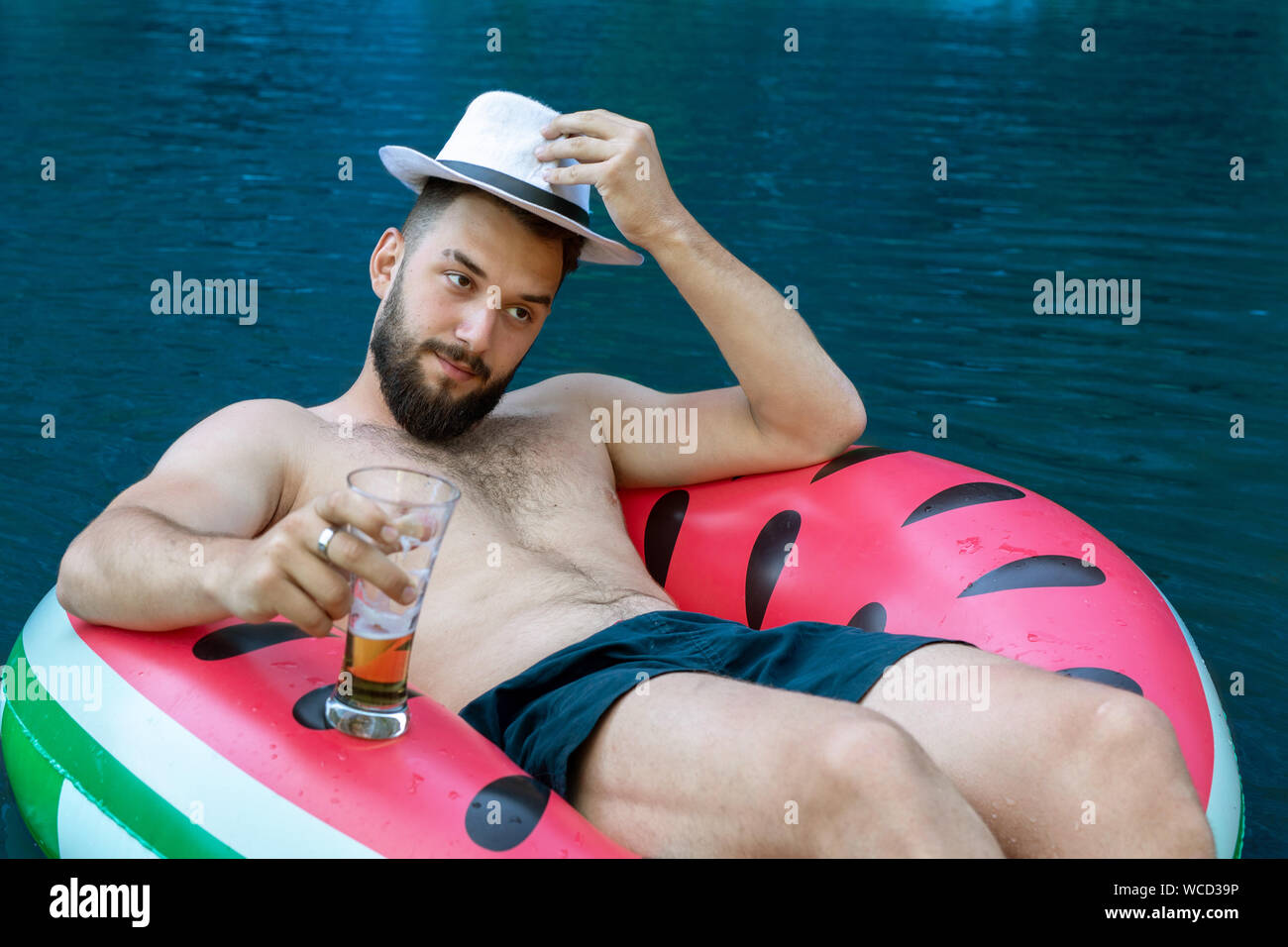 Giovane uomo attraente toglie il suo cappello, accoglie con favore. Nuota giacente in un cerchio gonfiabile con un bicchiere di birra Foto Stock