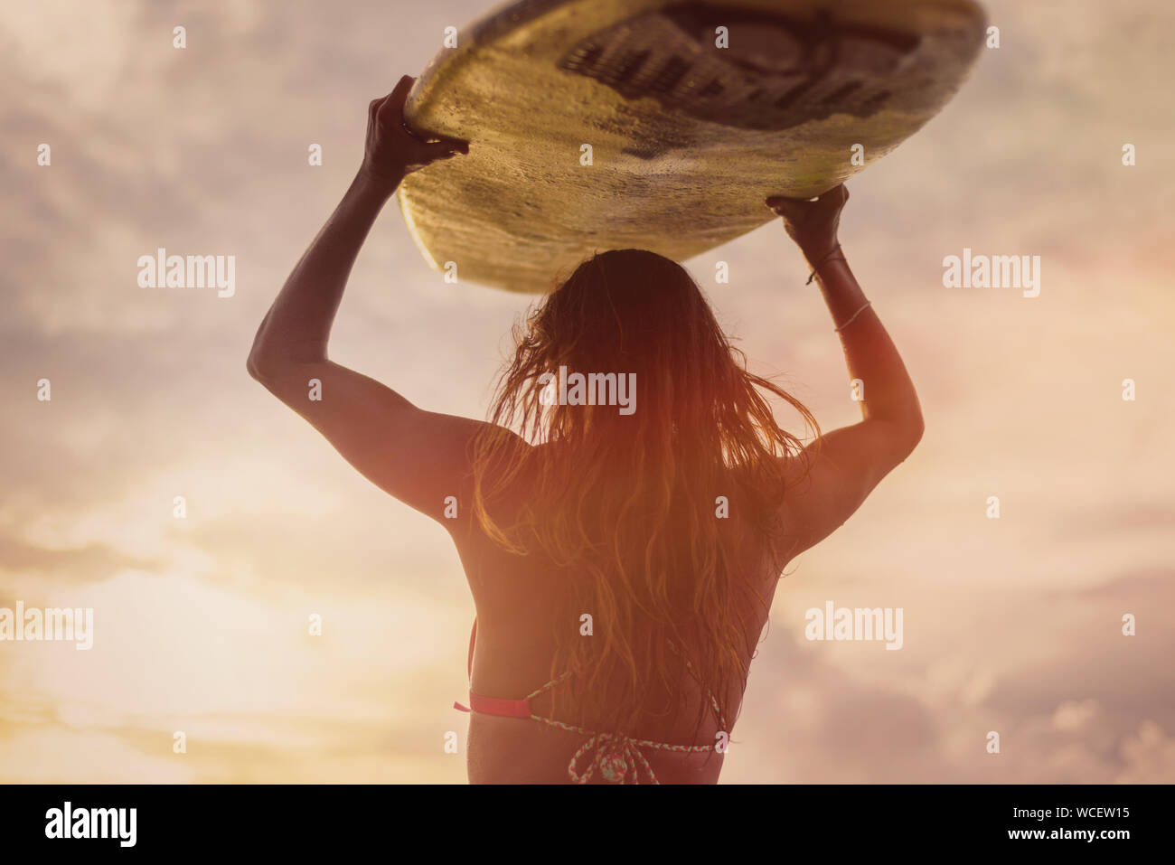 Un surfista con capelli lunghi in piedi indietro alla sua cella. Essa detiene un bianco con la tavola da surf sul suo capo al tramonto. Surfer e oceano. Colorazione calda. Medium Shot Foto Stock