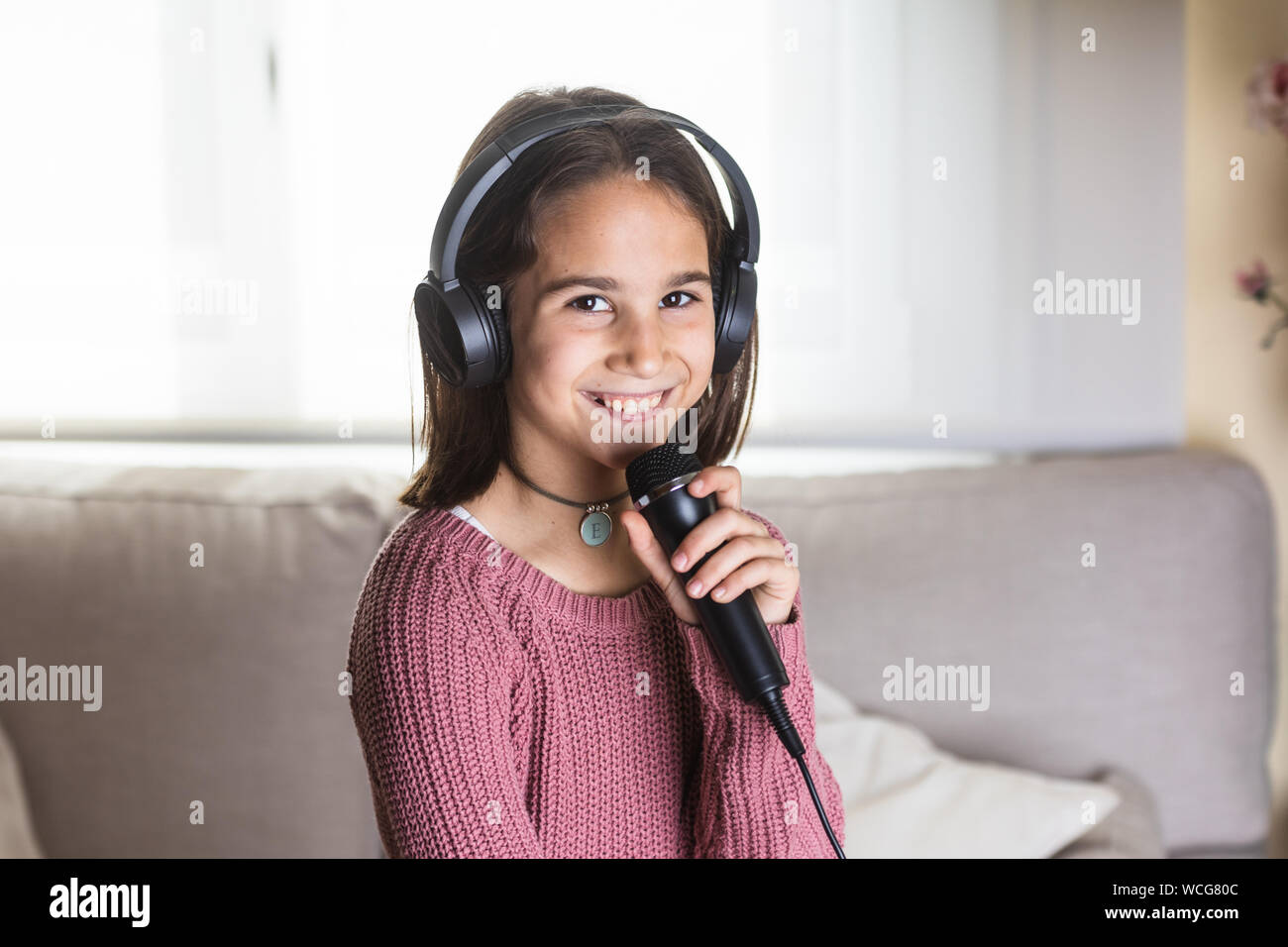 Niña cantando feliz con auriculares y micrófono en interior de casa Foto Stock