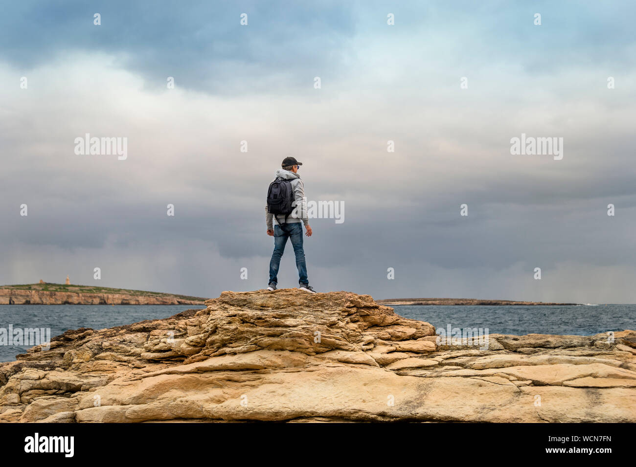 Uomo con zaino in piedi sulla parte superiore delle rocce che guarda al mare, moody sky. Foto Stock