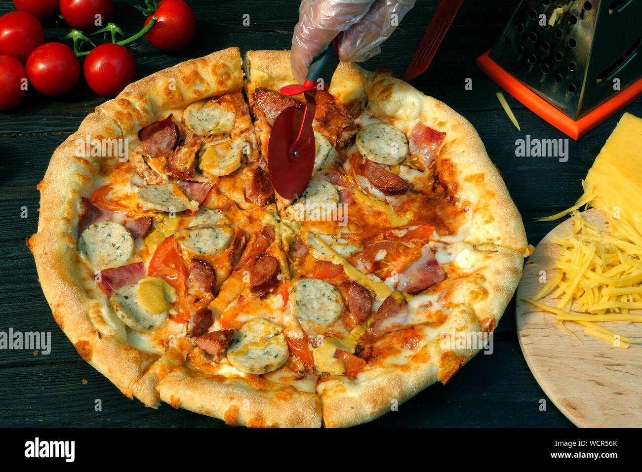 Tagliare la pizza, su fette, con salsiccia, pomodoro e formaggio, con uno speciale coltello - coltello ruota . Sul tavolo di legno vi sono anche i pomodori e grattugiato chee Foto Stock