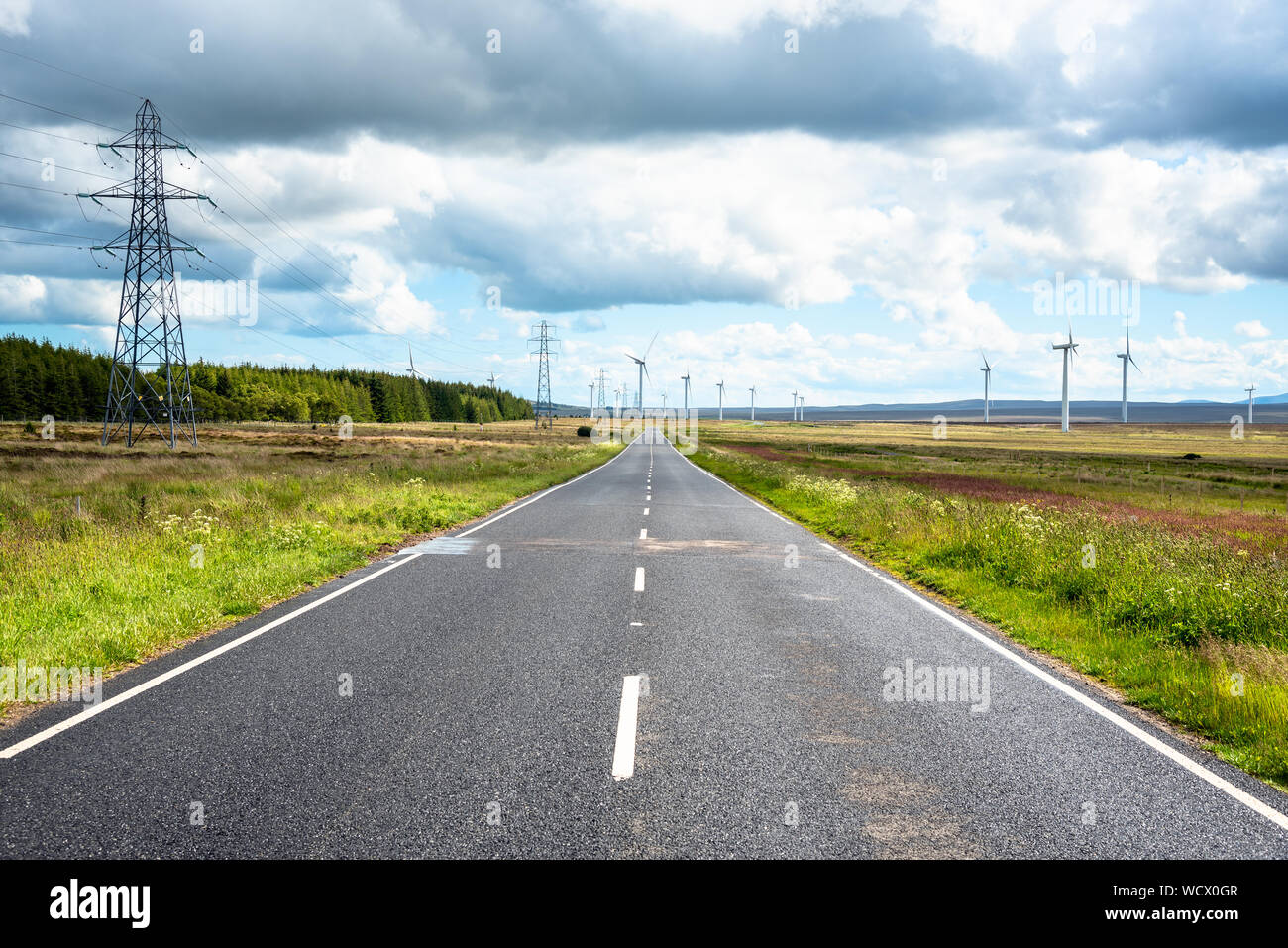 Strada deserta di una fattoria eolica su un nuvoloso giorno d'estate. Di energia elettrica ad alta tensione linea di tralicci la strada. Foto Stock