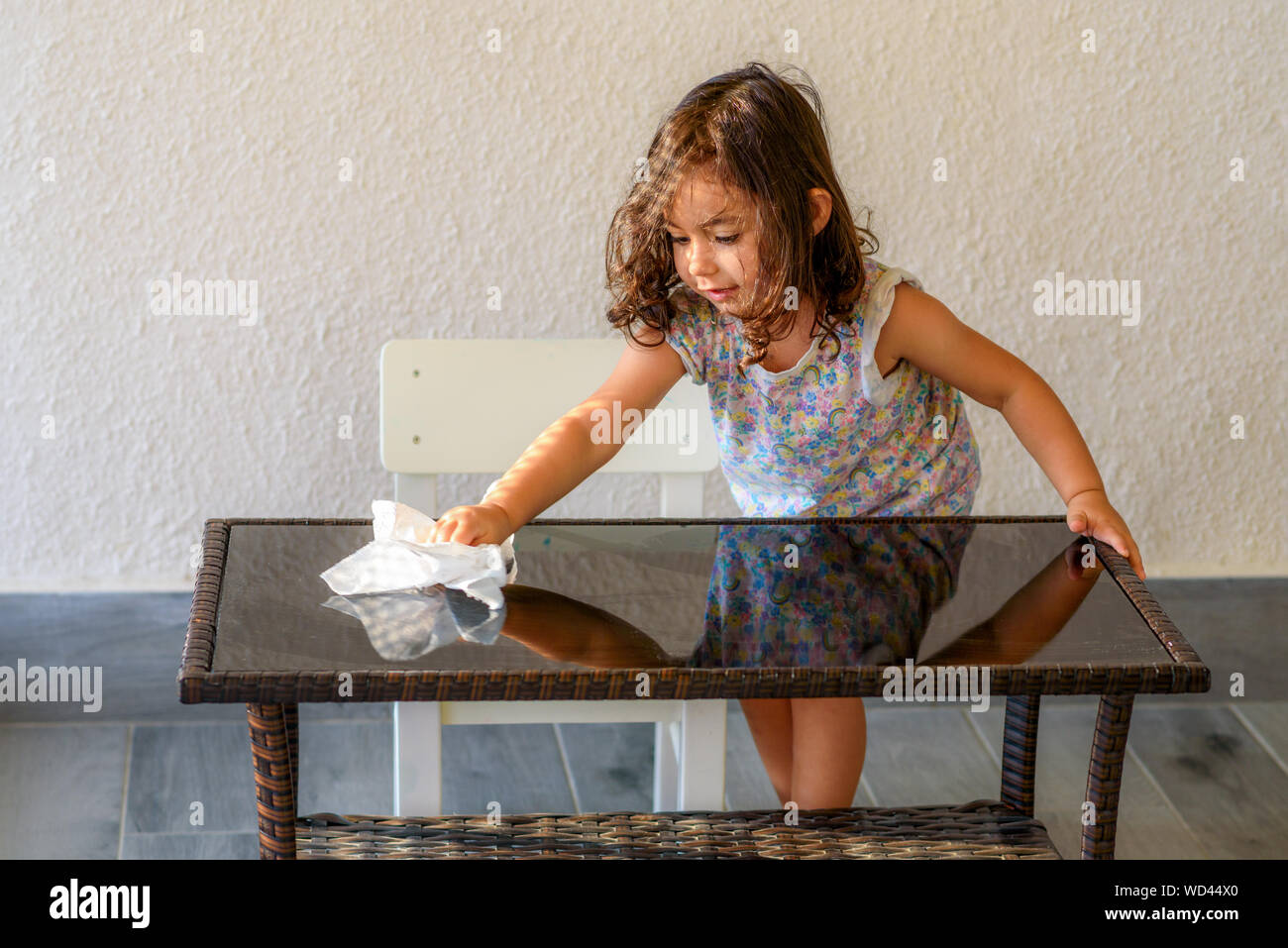 Carino brunette bambino aiuta la sua mamma lavaggio design moderno mobili da giardino, tavolo di vetro sulla terrazza di casa. Bambina rendono la pulizia con acqua e una soluzione detergente spray. Foto Stock