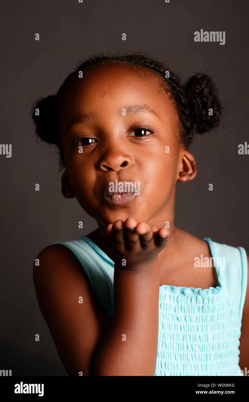 African bambina soffia un bacio con una luce blu sul vestito e capelli intrecciati, isolata su uno sfondo nero. Foto Stock