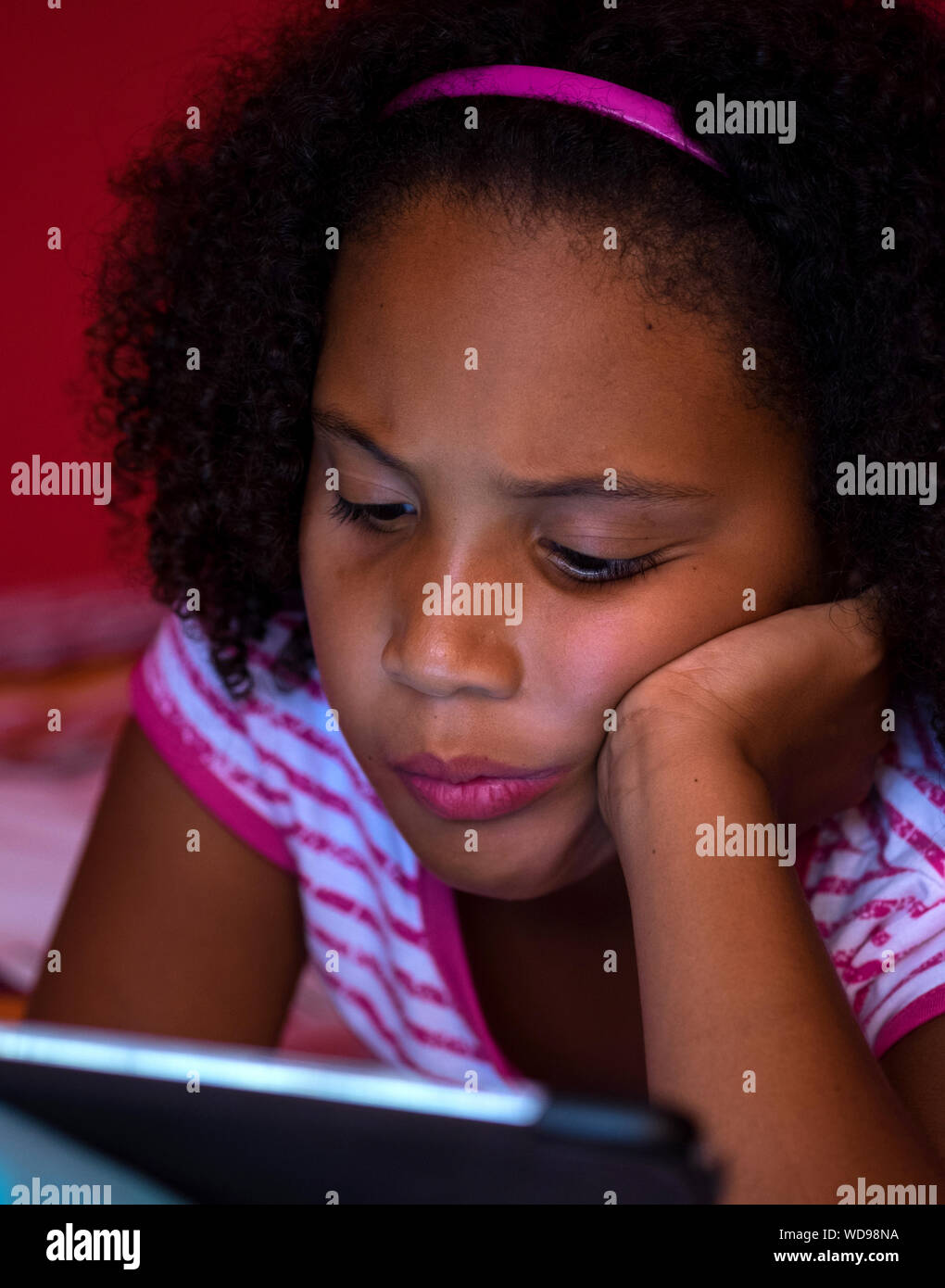 Bambino in profonda concentrazione con una cattiva postura, giocando sul suo tablet sul suo letto, indossando una striata rosa e bianca t-shirt. Foto Stock