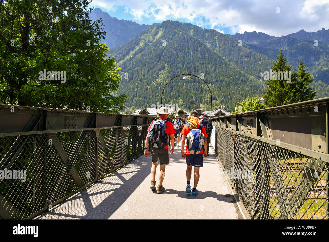 Turisti ed escursionisti attraversando un ponte pedonale sopra la linea ferroviaria con le montagne sullo sfondo in estate, Chamonix-Mont-Blanc, alpi, Francia Foto Stock
