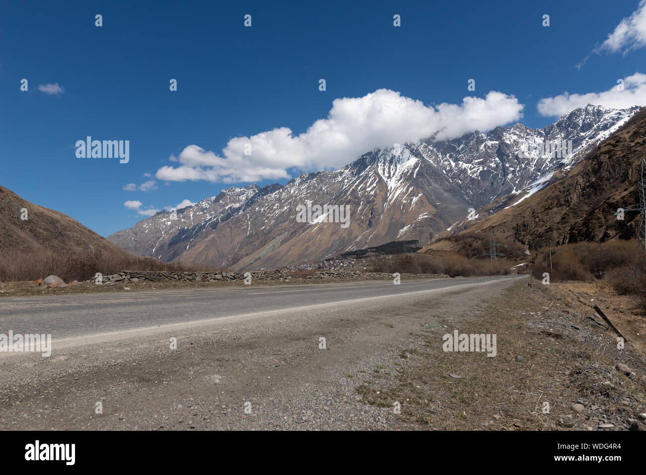 La Georgia, strada militare in primavera con la montagna, il ghiacciaio e un piccolo villaggio in vista, cielo blu Foto Stock