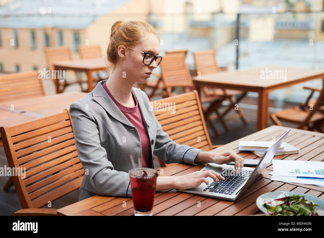 Dai capelli rossi imprenditrice gravi in occhiali lavora come libero professionista ha la seduta e la digitazione sul computer portatile durante il pranzo di affari in outdoor cafe Foto Stock
