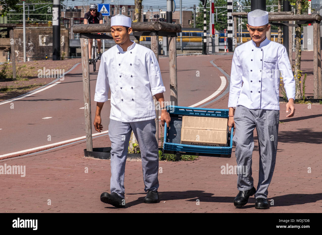 Amsterdam, Paesi Bassi - 30 Giugno 2019: due maschi giovani aiutanti di cucina in uniforme blu portano cassa con carne di agnello box lungo la strada nei pressi IJdok. Foto Stock