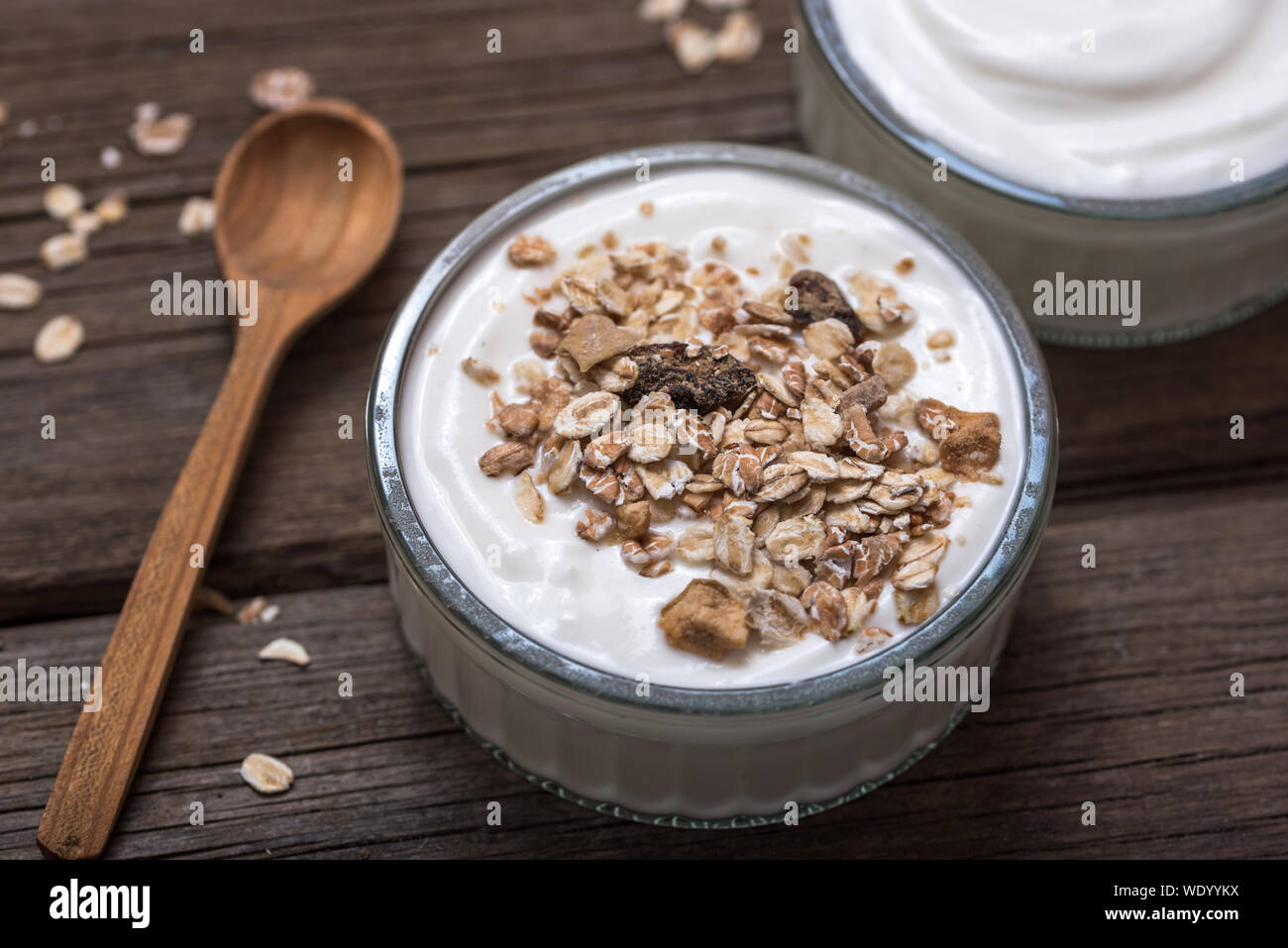 Yogurt bianco in recipiente di vetro su una vecchia scrivania in legno con il vecchio cucchiaio di legno e farina di avena sulla parte superiore. Foto Stock