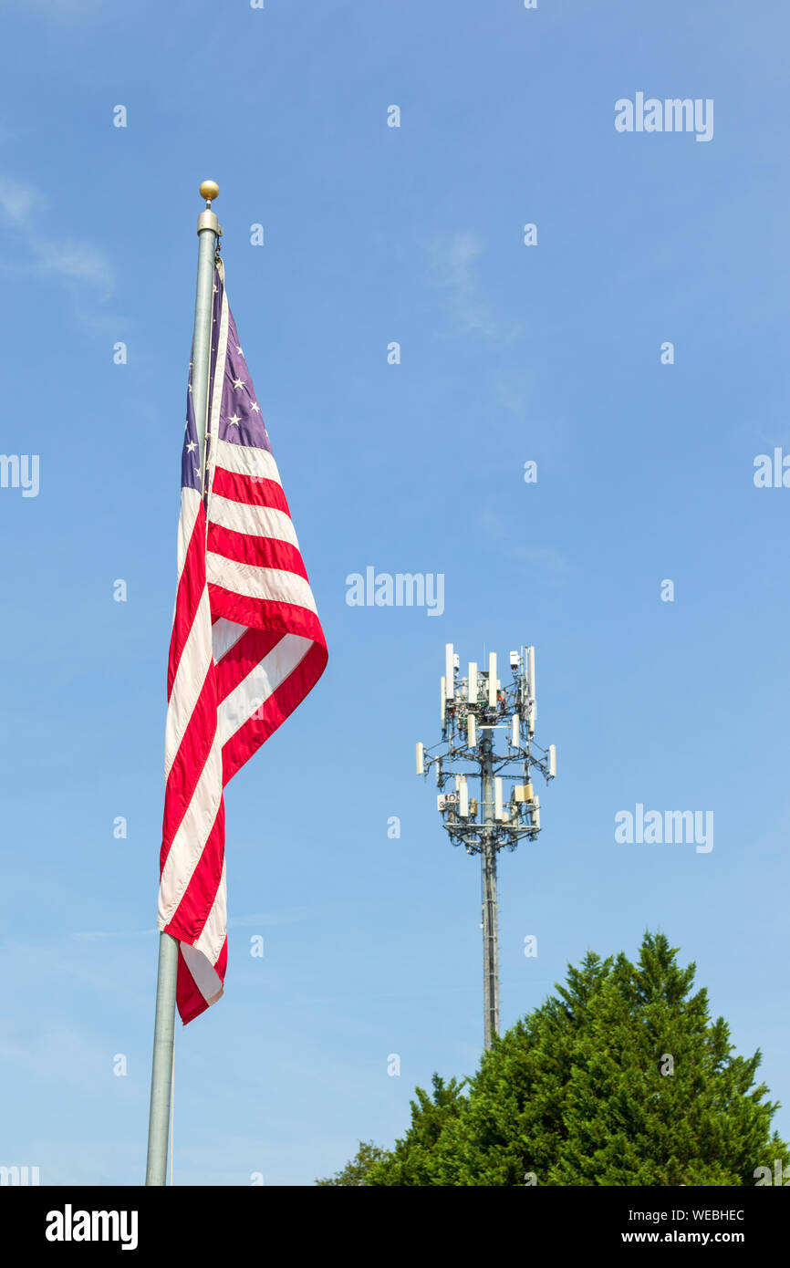 Una bandiera americana sulla bandiera pole affiancato ad un telefono cellulare la torre e il picco di un albero sempreverde. Cielo blu sullo sfondo. Copia dello spazio. Foto Stock