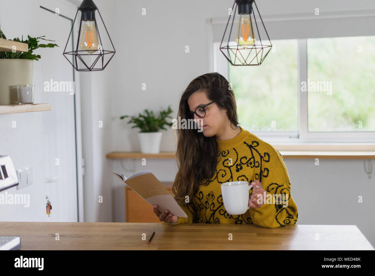 Donna con gli occhiali tenendo una tazza e la lettura da un notebook su un banco di lavoro in legno all'interno di una casa. Foto Stock