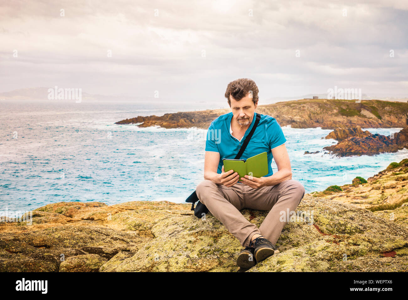 Uomo seduto all'aperto sulla scogliera la lettura di un e-book da mare Foto Stock