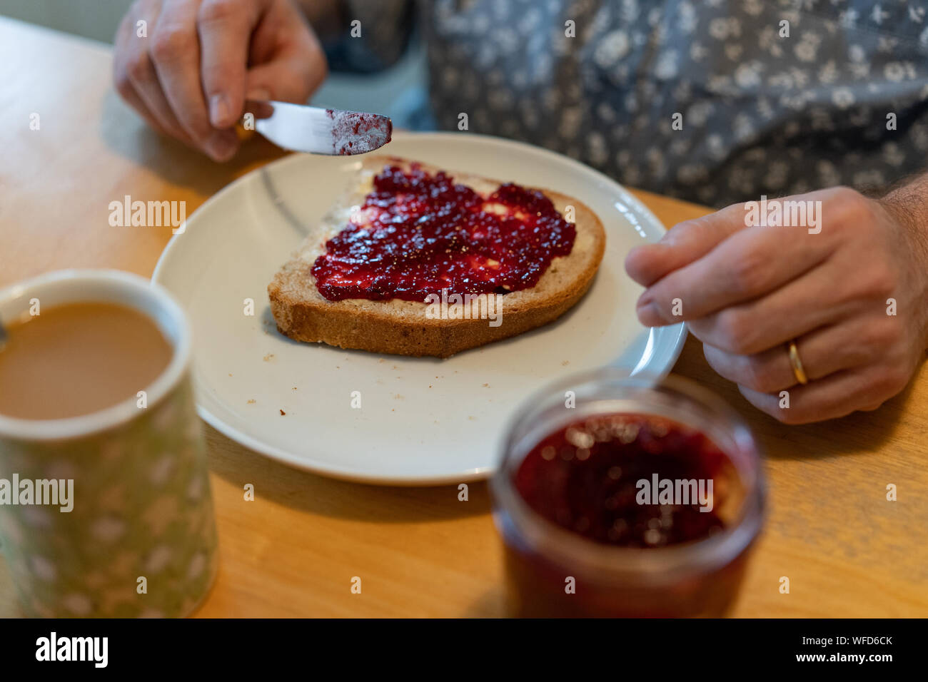 Un uomo seduto in cucina e la diffusione della gelatina sul suo pane con caffè e un bicchiere di gelatina sul tavolo di betulla. Foto Stock