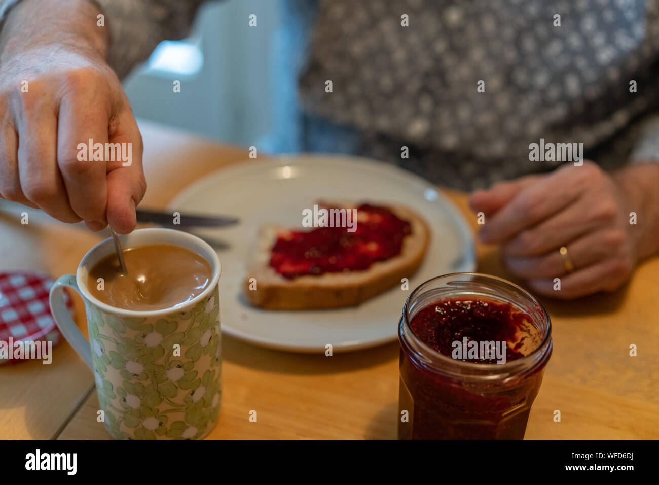 Un uomo seduto in cucina suscita il suo caffè, con pane sulla sua piastra e un bicchiere di gelatina sul tavolo di betulla Foto Stock