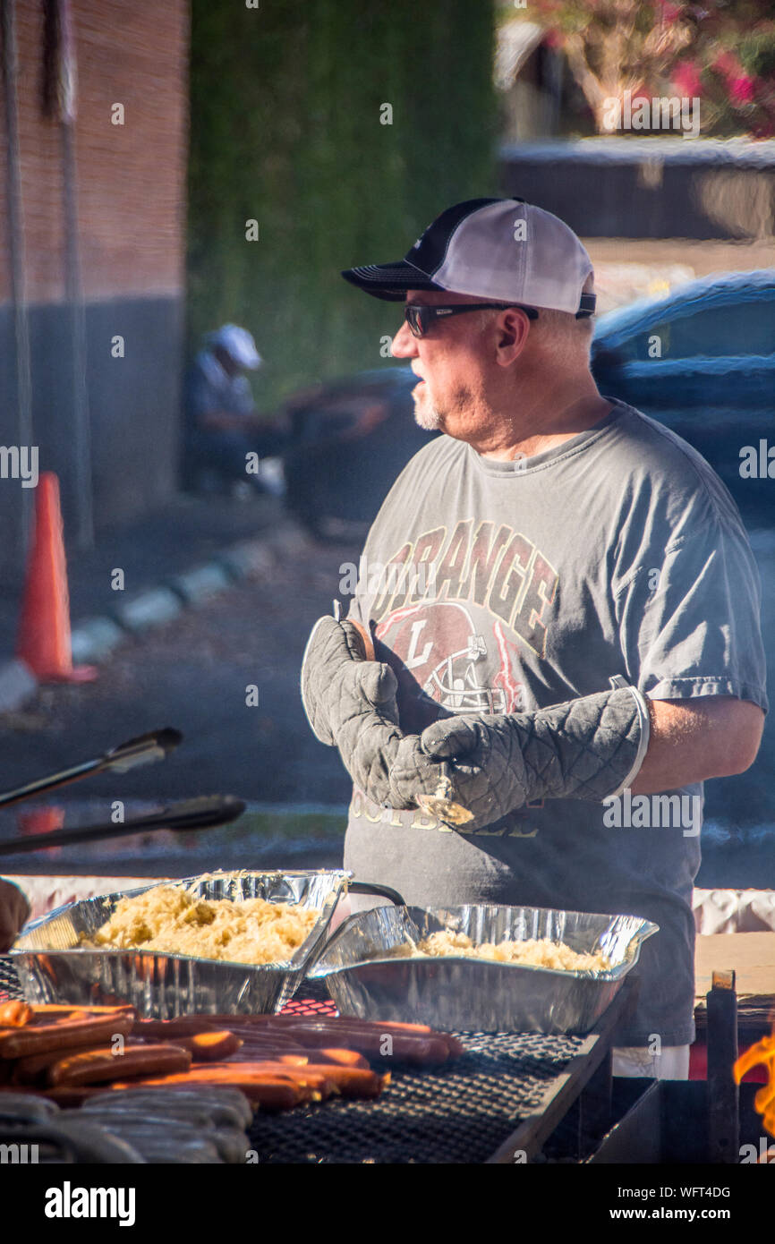 Un senior che indossa guanti, si ferma per riposare, tra la cottura di alcuni hot dog. Foto Stock