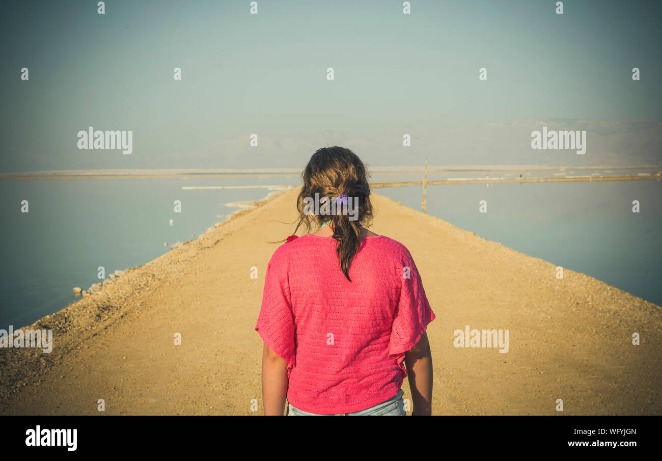 Vista posteriore di una ragazza nella parte superiore rossa camminando sul marciapiede in mezzo al mare contro Sky Foto Stock