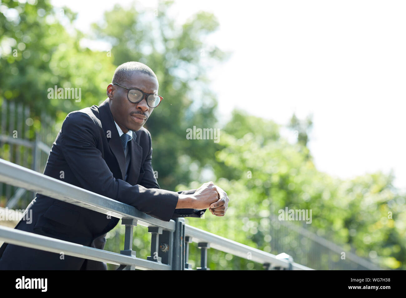 Ritratto di elegante uomo afro-americano di indossare tuta e bicchieri in posa appoggiata sulla ringhiera all'aperto nel parco, spazio di copia Foto Stock