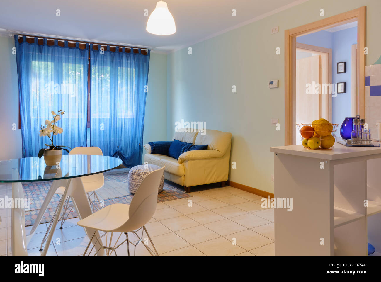 Elegante soggiorno di un piccolo appartamento con un round moderno tavolo in vetro e una parte di angolo cottura visibile sul lato destro Foto Stock