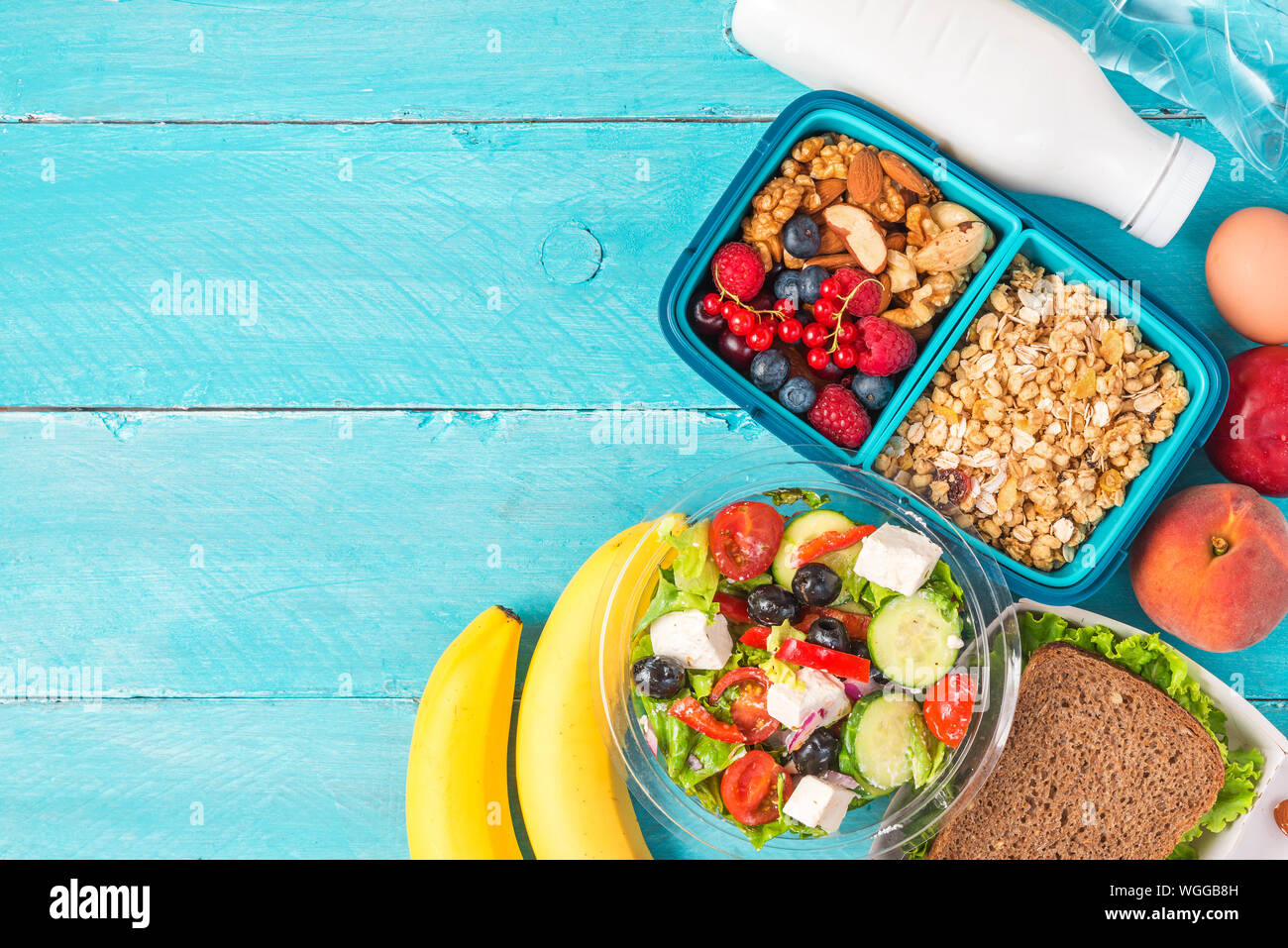 Set di pranzo sano di scatole con insalata greca, sandwich, granola, verdure, dadi, bacche, banana, uova, pesche e yogurt su sfondo blu. top vi Foto Stock