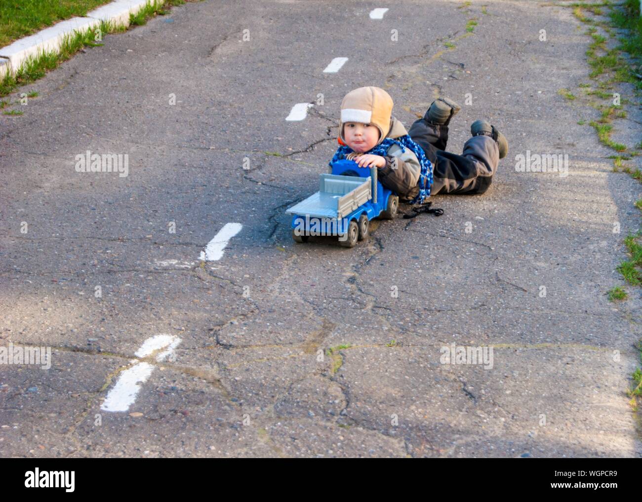 Un piccolo ragazzo di tre anni è sdraiato sul pavimento vicino alla scuola materna con il suo giocattolo carrello - molla, può raffreddare vestiti caldi, giochi all'aperto, un bambino in un cappello e una giacca Foto Stock