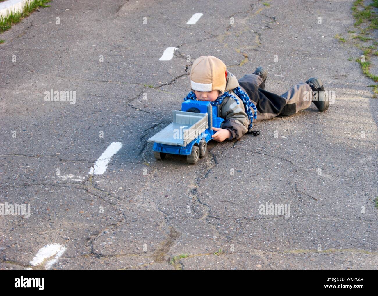 Un piccolo ragazzo di tre anni è sdraiato sul pavimento vicino alla scuola materna con il suo giocattolo carrello - molla, può raffreddare vestiti caldi, giochi all'aperto, un bambino in un cappello e una giacca Foto Stock