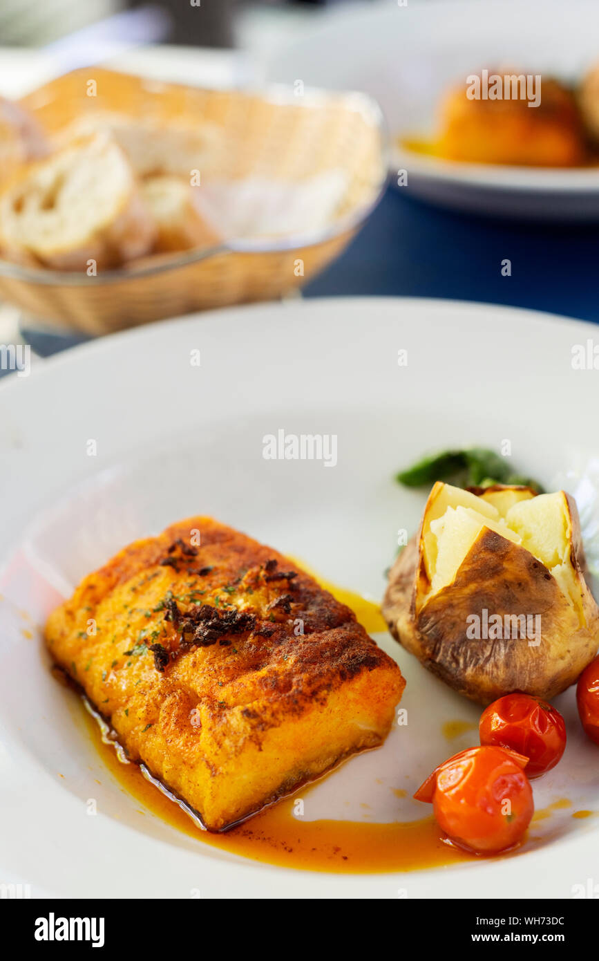 Primo piano di un pezzo di cotto martoriata il merluzzo in un piatto di portata in ceramica bianca, guarnita con patate arrosto, alcuni fritti pomodori ciliegia e qualche fritto verde p Foto Stock
