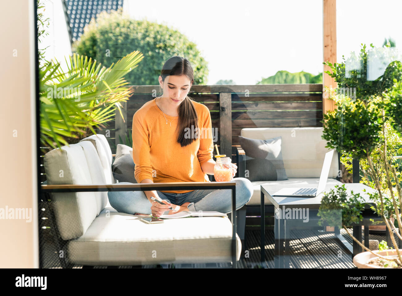 Giovane donna seduta sul lettino sulla terrazza con bibita salutare e notebook Foto Stock