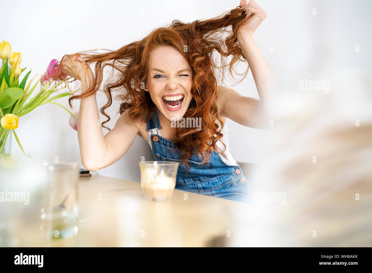 Ritratto di donna allegra tira i capelli mentre è seduto con bevanda a tavola in camera Foto Stock