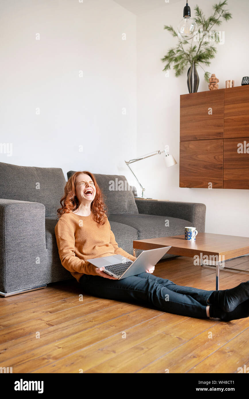 Lunghezza completa di allegra donna ridere durante la seduta con il computer portatile nel salotto di casa Foto Stock