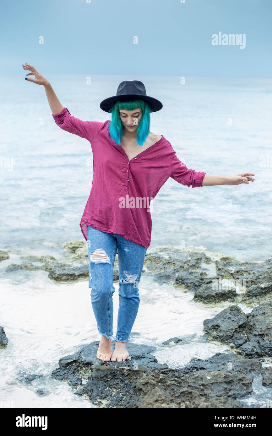 Giovane donna con tinti di blu e verde di bilanciamento dei capelli a piedi nudi sulla roccia di fronte al mare, Spagna Foto Stock