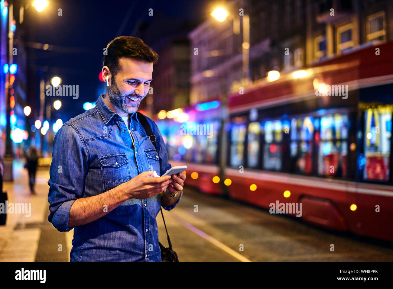 Uomo sorridente con cuffie wireless utilizza lo smartphone durante l'attesa per il tram di notte Foto Stock