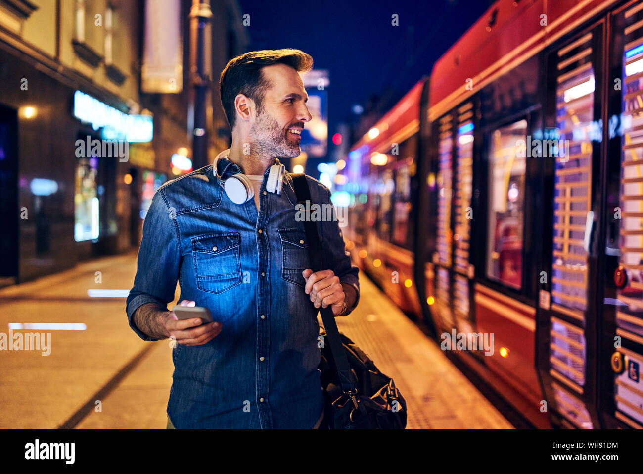 Uomo sorridente con il suo smartphone nella città di notte mentre si attende per il tram Foto Stock
