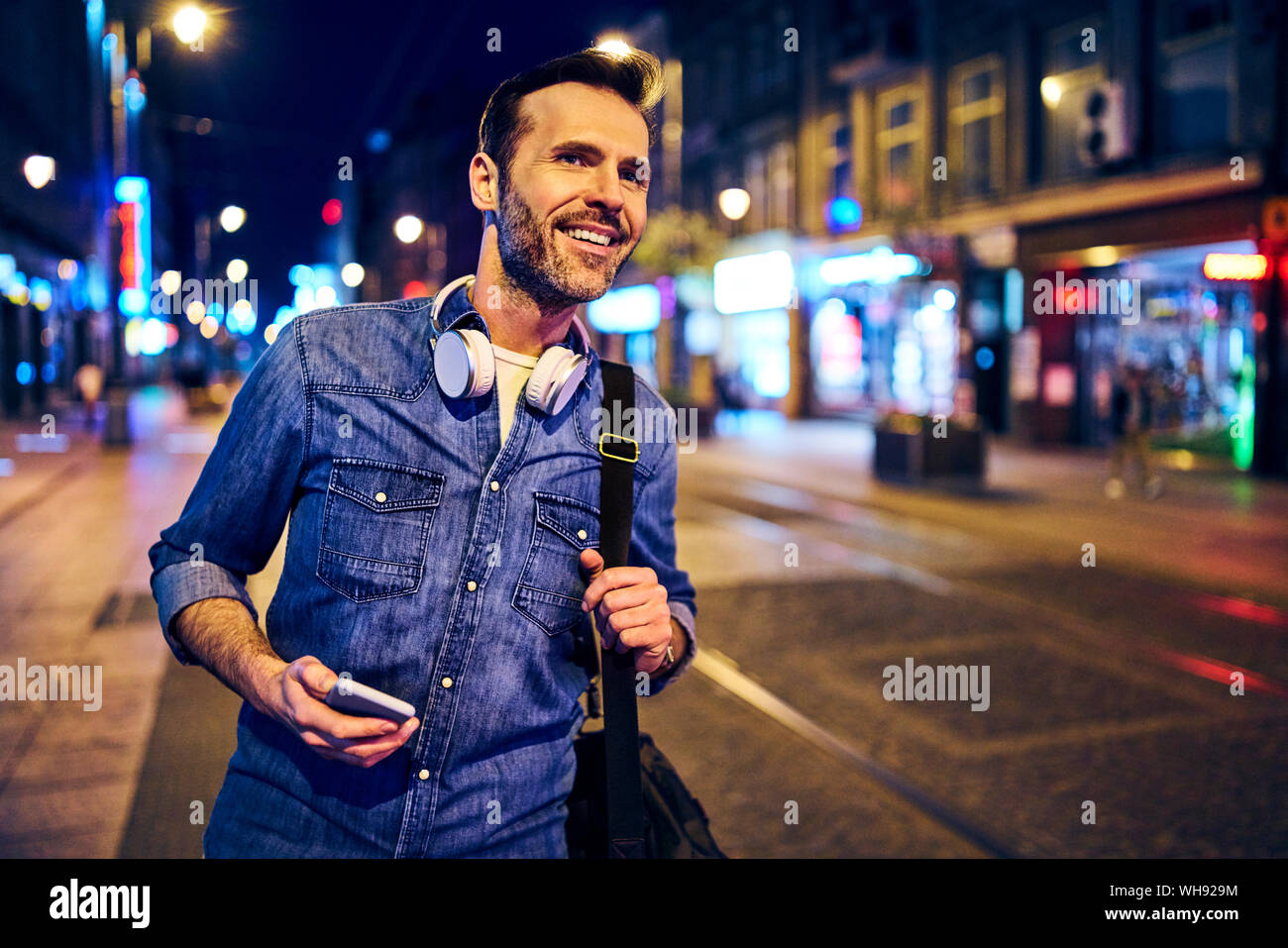 Sorridente con lo smartphone nella città di notte mentre si attende per il tram Foto Stock