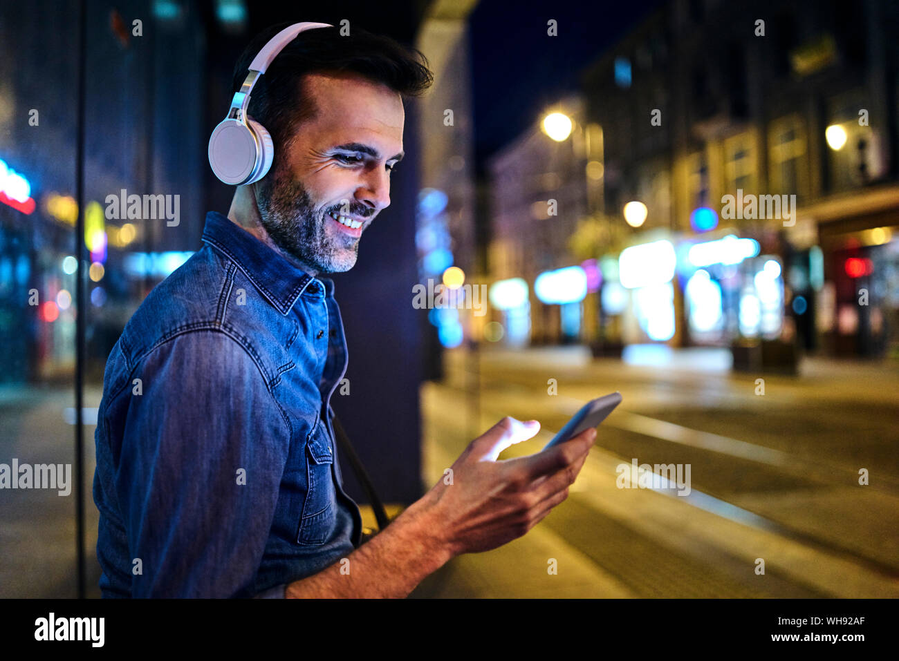Uomo sorridente con cuffie utilizza lo smartphone mentre si è in attesa per il bus notturno in città Foto Stock