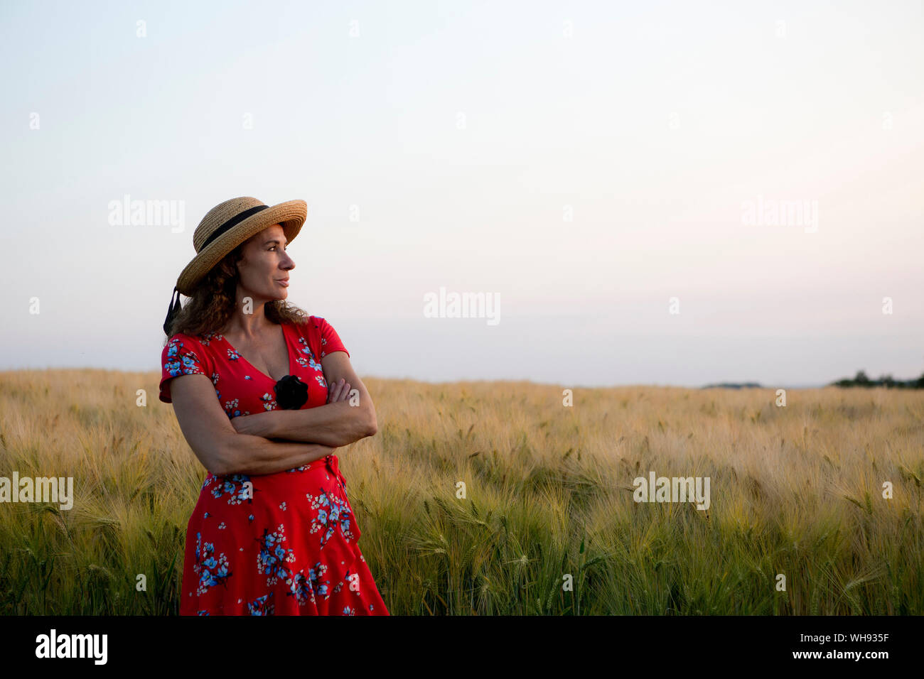 Donna che indossa cappello di paglia e vestito estivo con disegno floreale in piedi nella parte anteriore del campo di grano Foto Stock