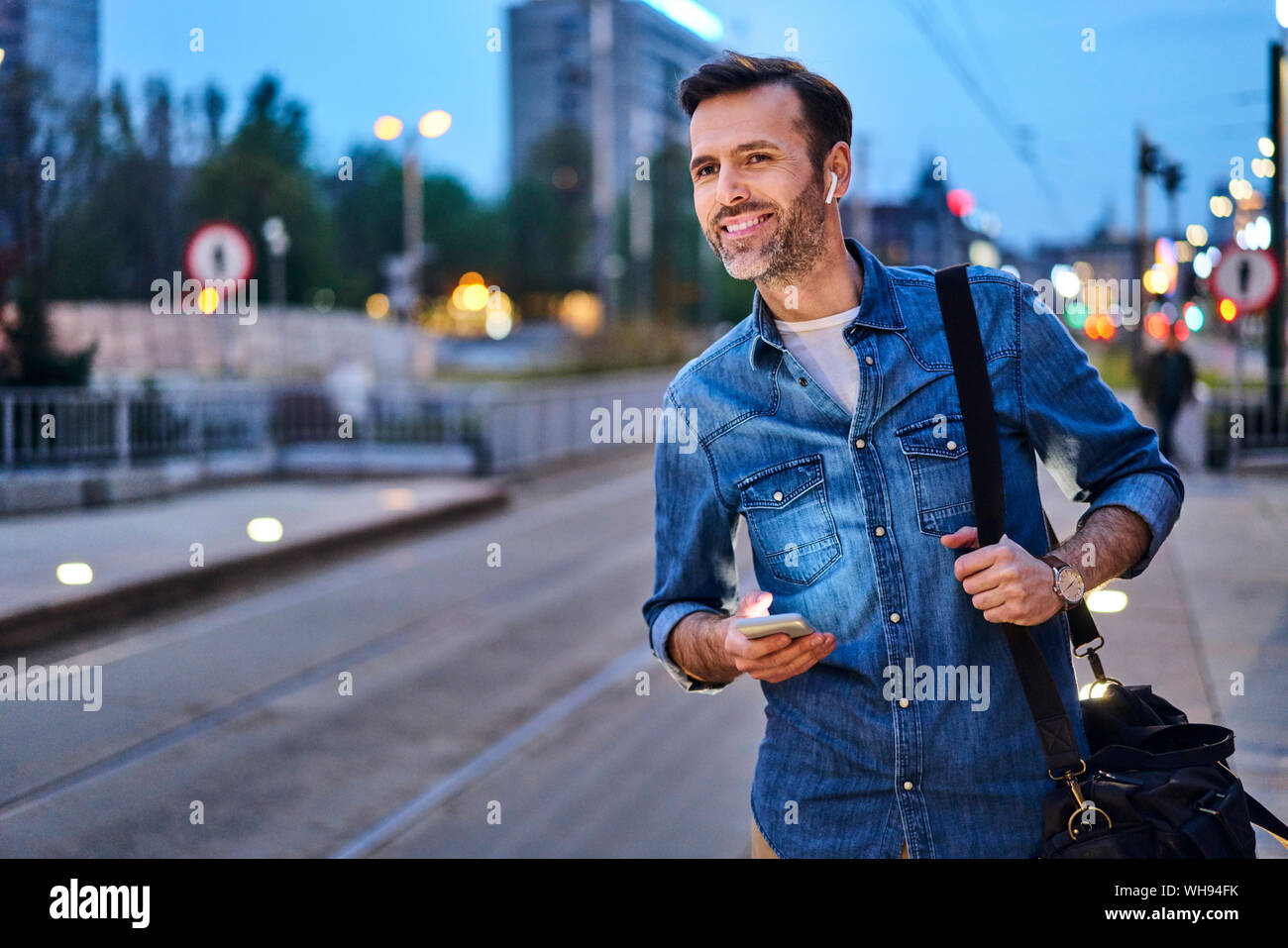 Uomo sorridente con cuffie wireless e lo smartphone in attesa alla fermata del tram durante spostamenti di sera dopo il lavoro Foto Stock