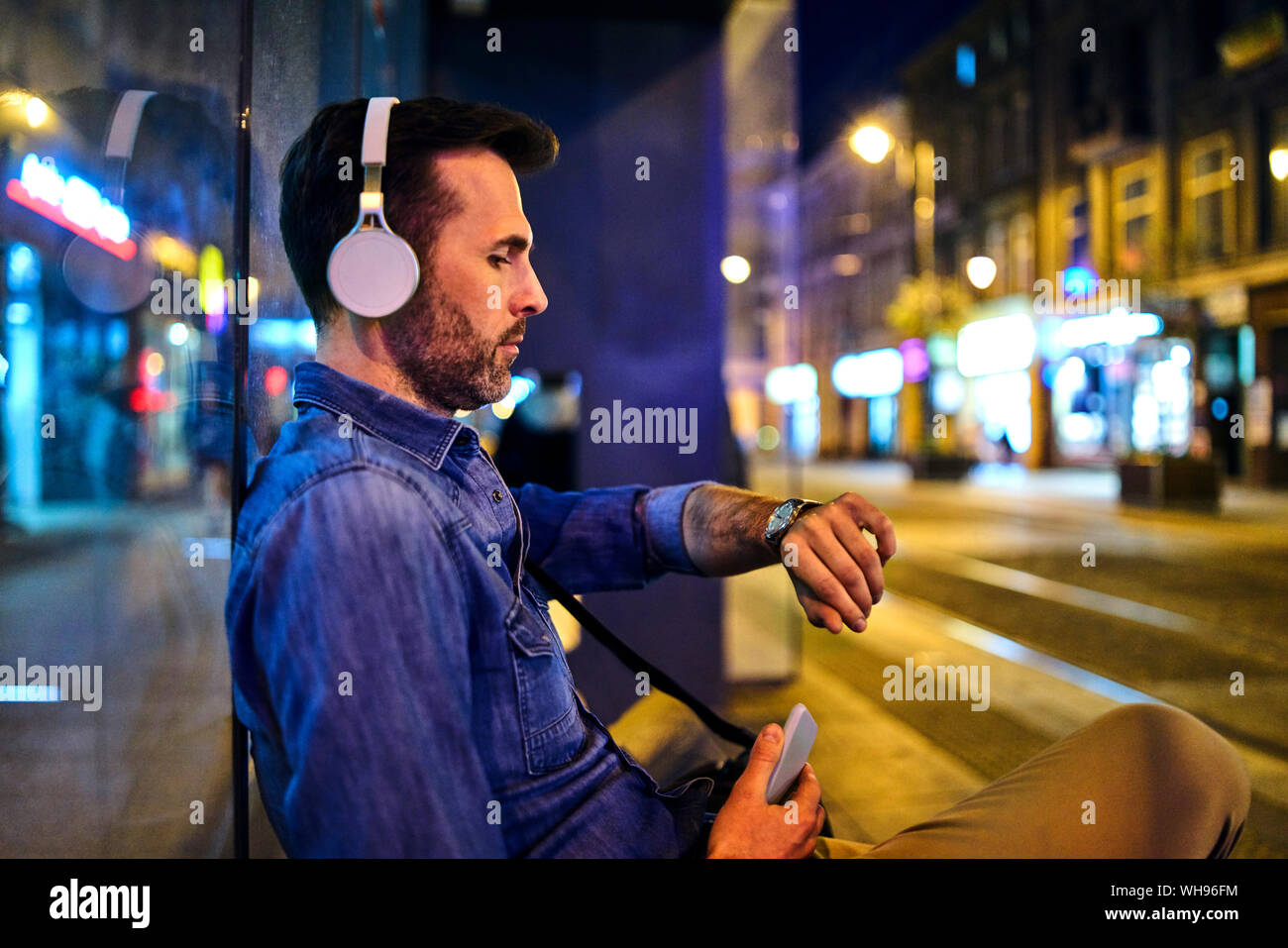 Uomo serio con cuffie wireless in attesa del bus notturno in città e controllando il tempo Foto Stock
