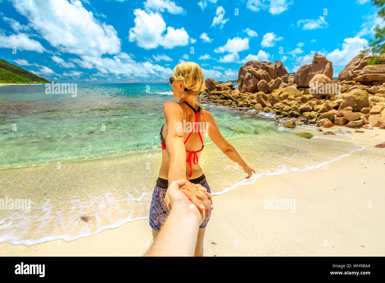 Bionda donna turistica in bikini tenendo la mano del partner ad Anse Caimano spiaggia con piscina naturale di La Digue, Seychelles, Oceano indiano, Africa Foto Stock