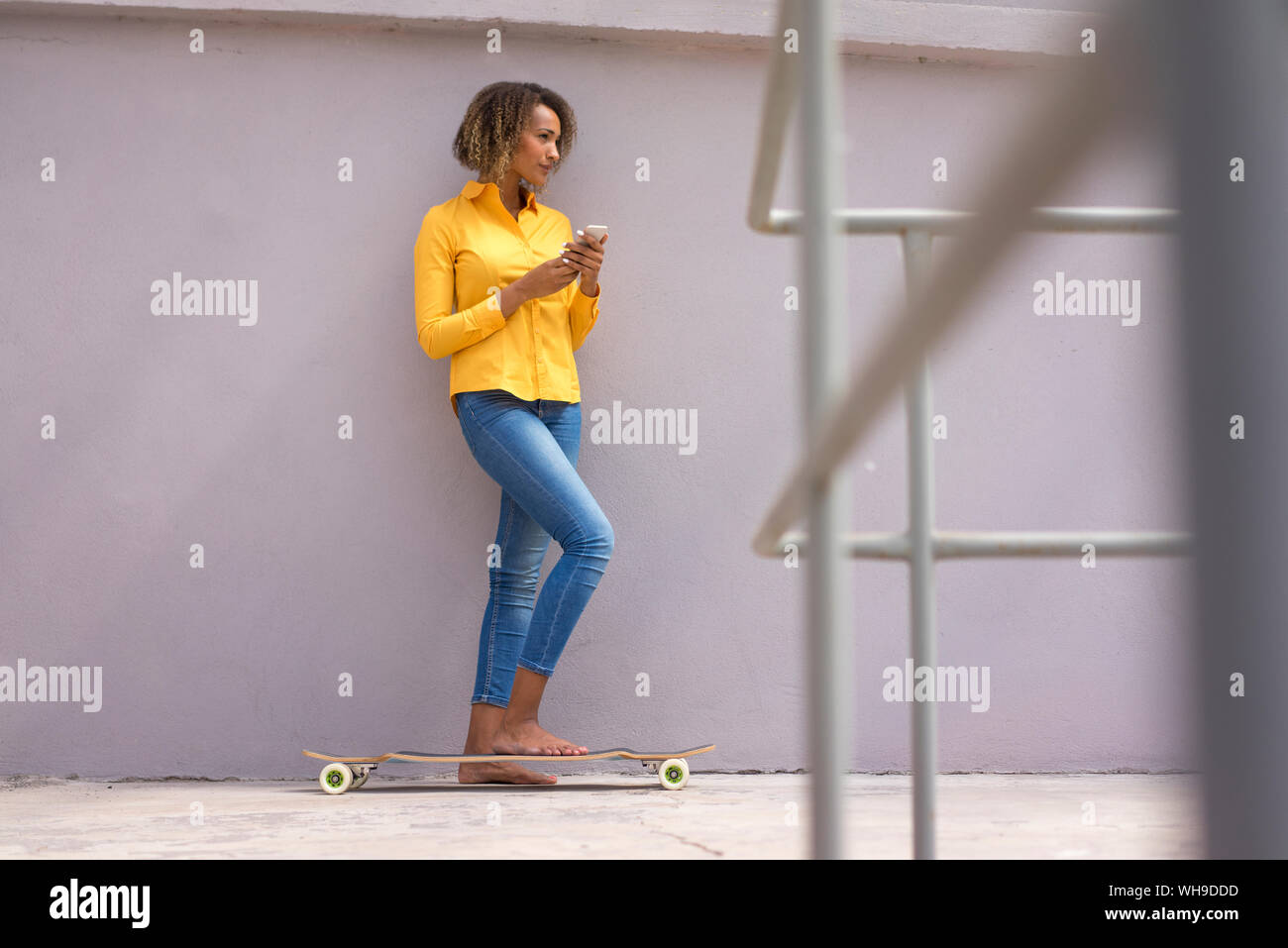 Giovane donna a piedi nudi con lo smartphone e lo skateboard indossare magliette di colore giallo e jeans Foto Stock