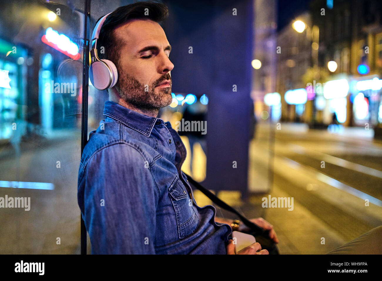 Uomo con cuffie wireless snoozing durante l'attesa per il bus notturno in città Foto Stock