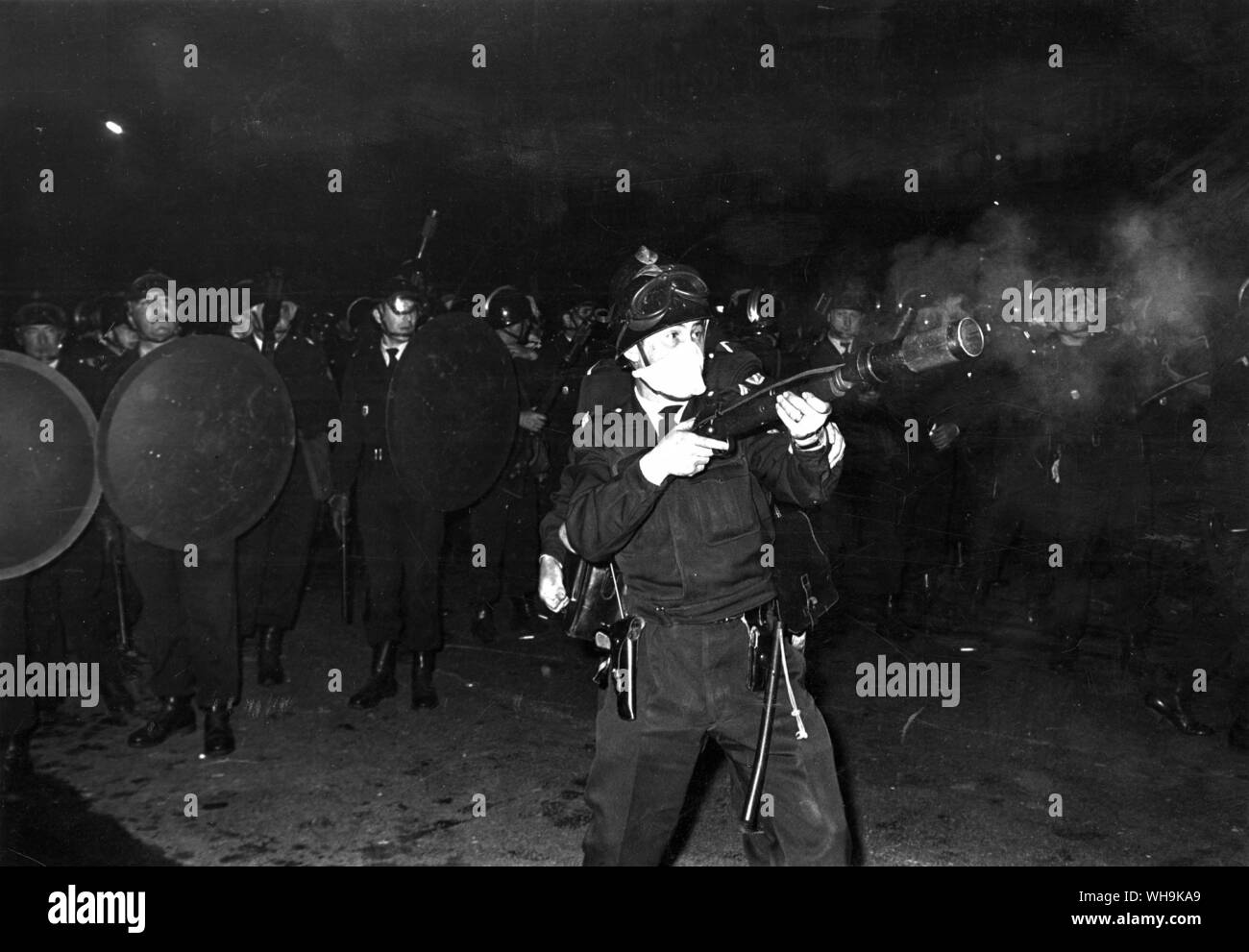 1968: Parigi scontri. Un uomo di polizia spara un gas lacrimogeni bomba in mezzo agli studenti, mentre i suoi compagni armati con scudi e bastoni preparare per la carica. Foto Stock
